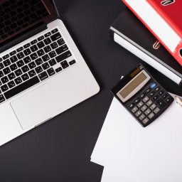 Biuro Usług Księgowych i Finansowych Anna Nowakowska - Laptop, kalkulator, dokumenty i segregatory na czarnym biurku. Widok z góry sugeruje pracę biurową, analizę danych lub przygotowywanie dokumentacji.