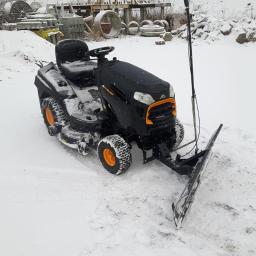 Bracia Wawrzyniak - Czarny traktorek do koszenia trawy z zamontowanym pługiem do śniegu, stoi na ośnieżonym terenie, w tle widoczne betonowe kręgi i elementy konstrukcyjne