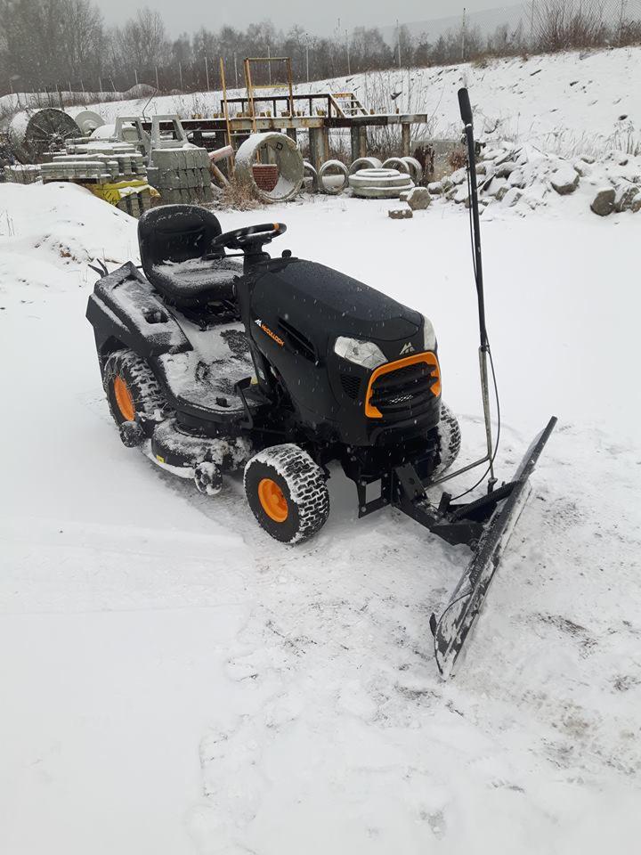 Czarny traktorek do koszenia trawy z zamontowanym pługiem do śniegu, stoi na ośnieżonym terenie, w tle widoczne betonowe kręgi i elementy konstrukcyjne