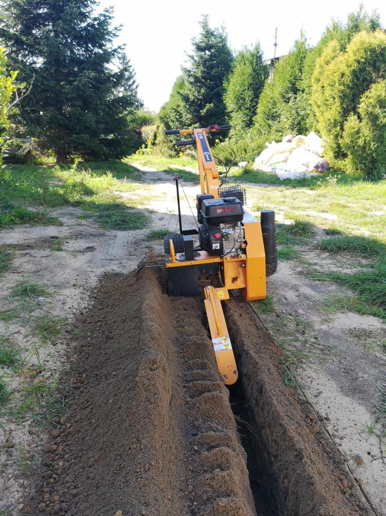 Żółta glebogryzarka spalinowa wykonuje wąski wykop w ziemi, widoczne świeżo wykopane rowy z grudami ziemi, w tle zielona trawa i drzewa.
