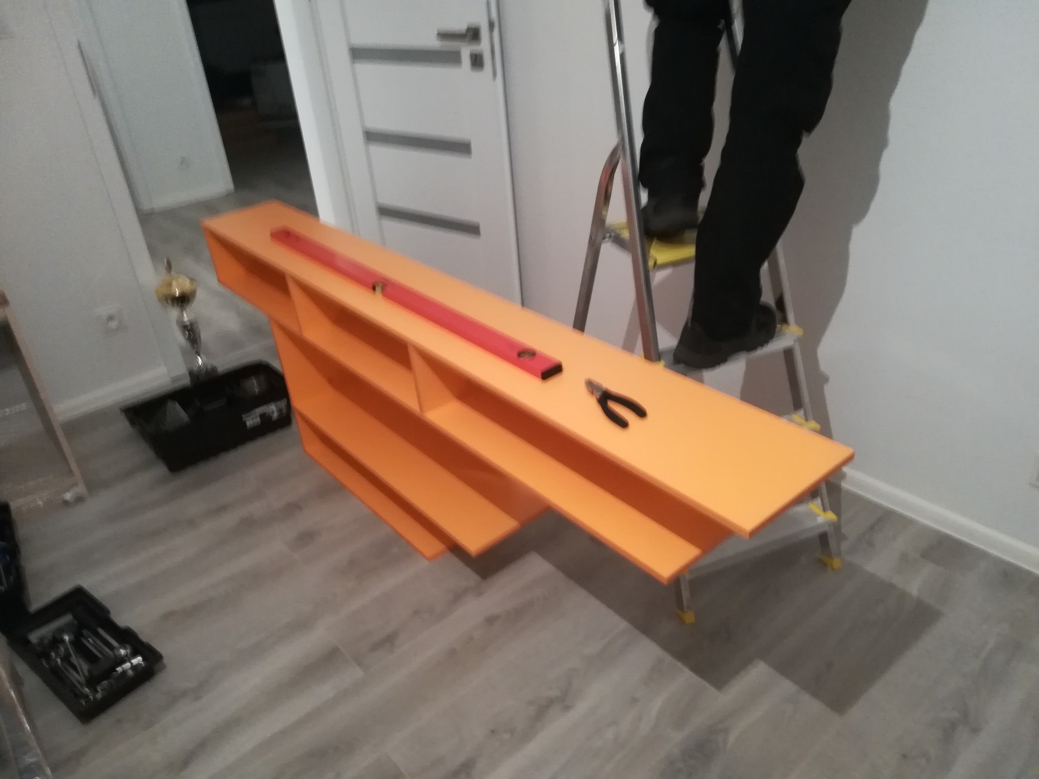 Pomarańczowa, wielopoziomowa półka z poziomnicą i kombinerkami na blacie, ustawiona w pomieszczeniu podczas montażu, z pracownikiem na drabinie w tle i skrzynką narzędziową na podłodze.