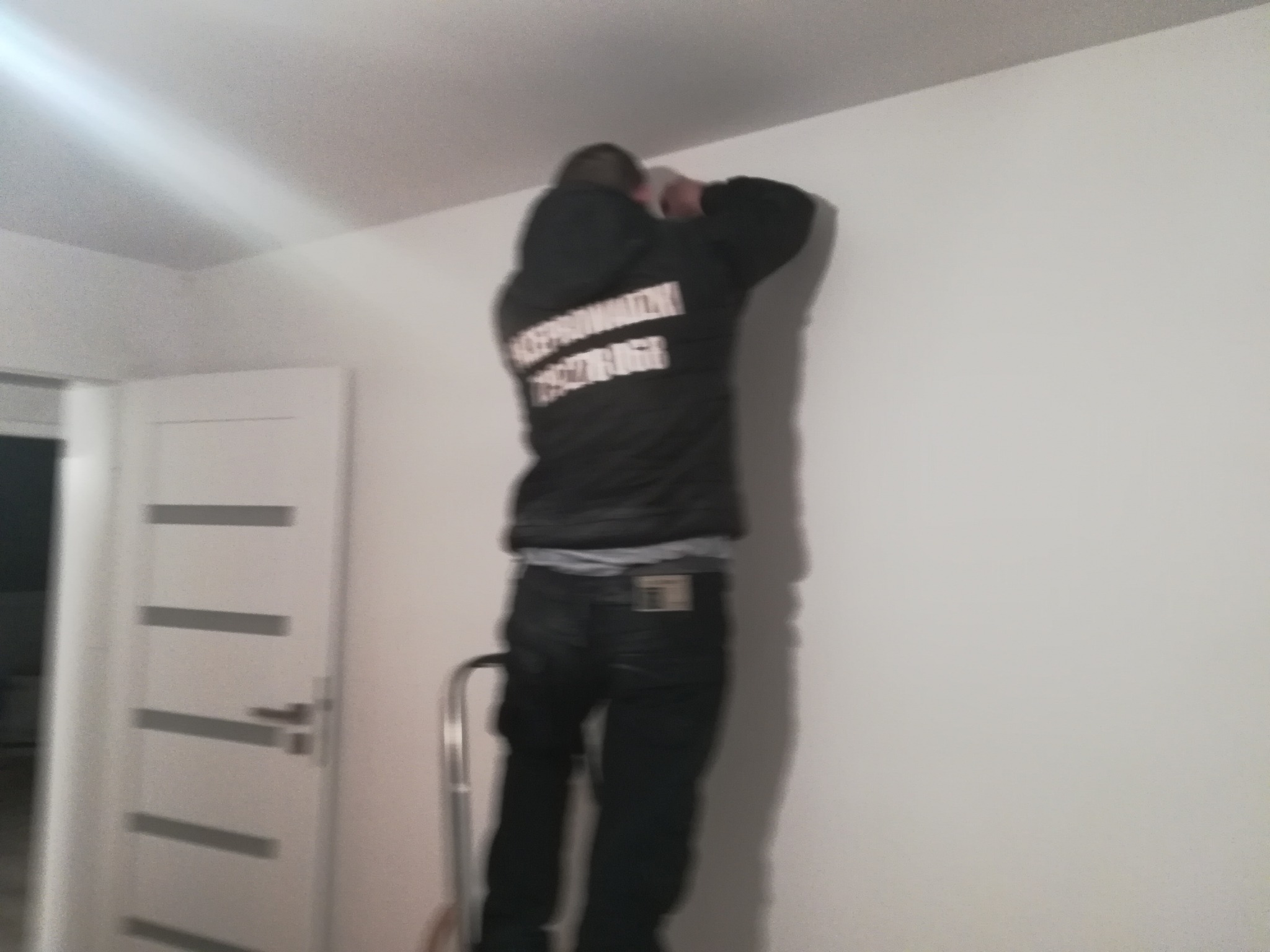 Mężczyzna w bluzie z napisem, stojący na drabince i mocujący coś do białej ściany w jasnym wnętrzu.