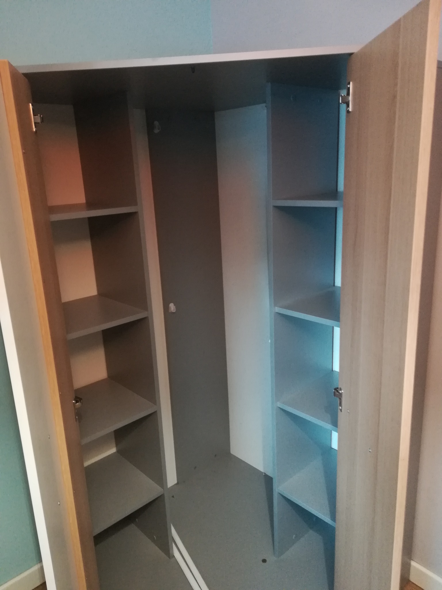 Wnętrze szafy narożnej z otwartymi drzwiami, widoczne półki i szary tył szafy, zamontowana w pokoju dziecięcym z błękitną ścianą.