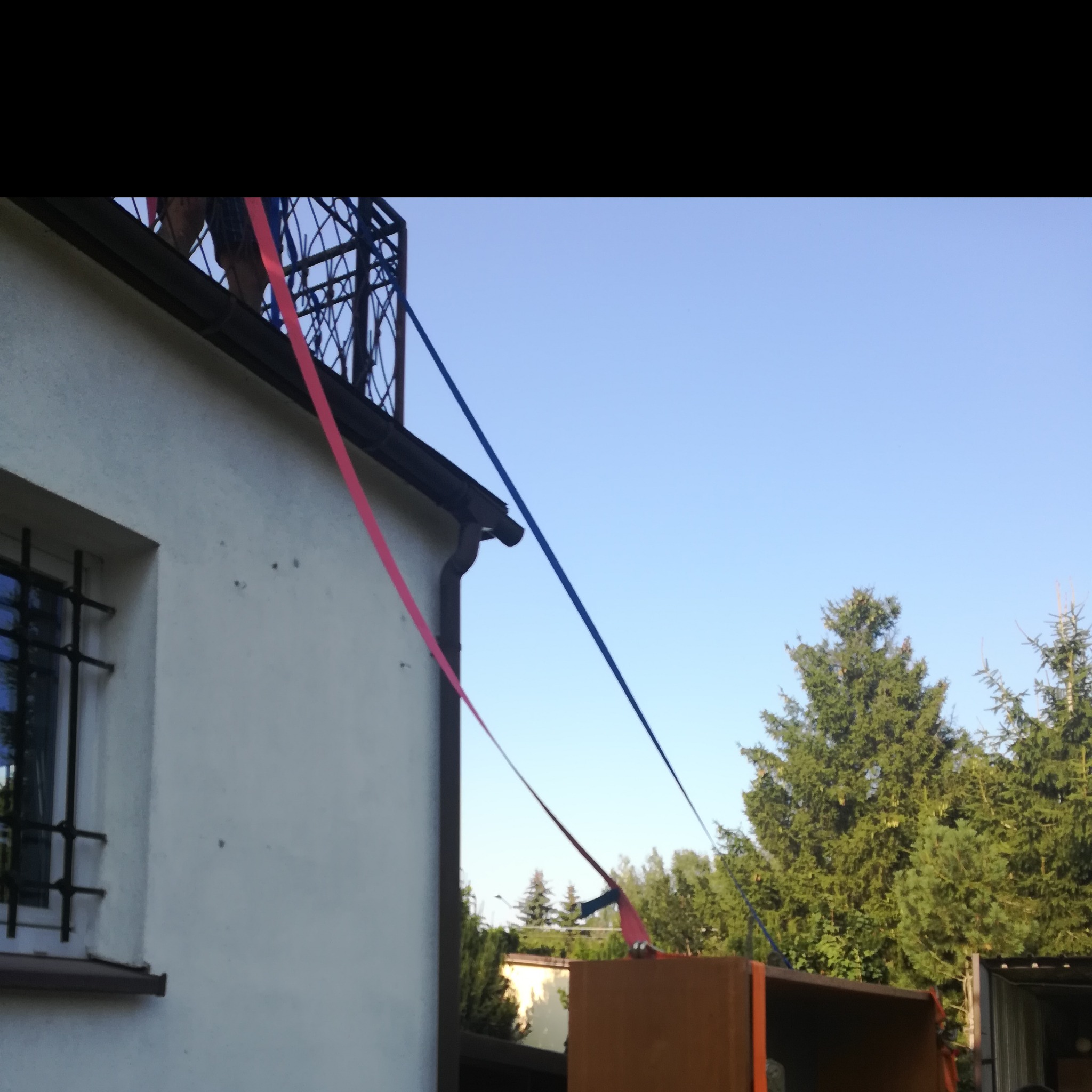 Regał wciągany na pasach przez balkon na pierwsze piętro, widok z dołu na linę czerwoną i niebieską przymocowaną do regału i balkonu budynku z białymi ścianami i oknem z kratami.