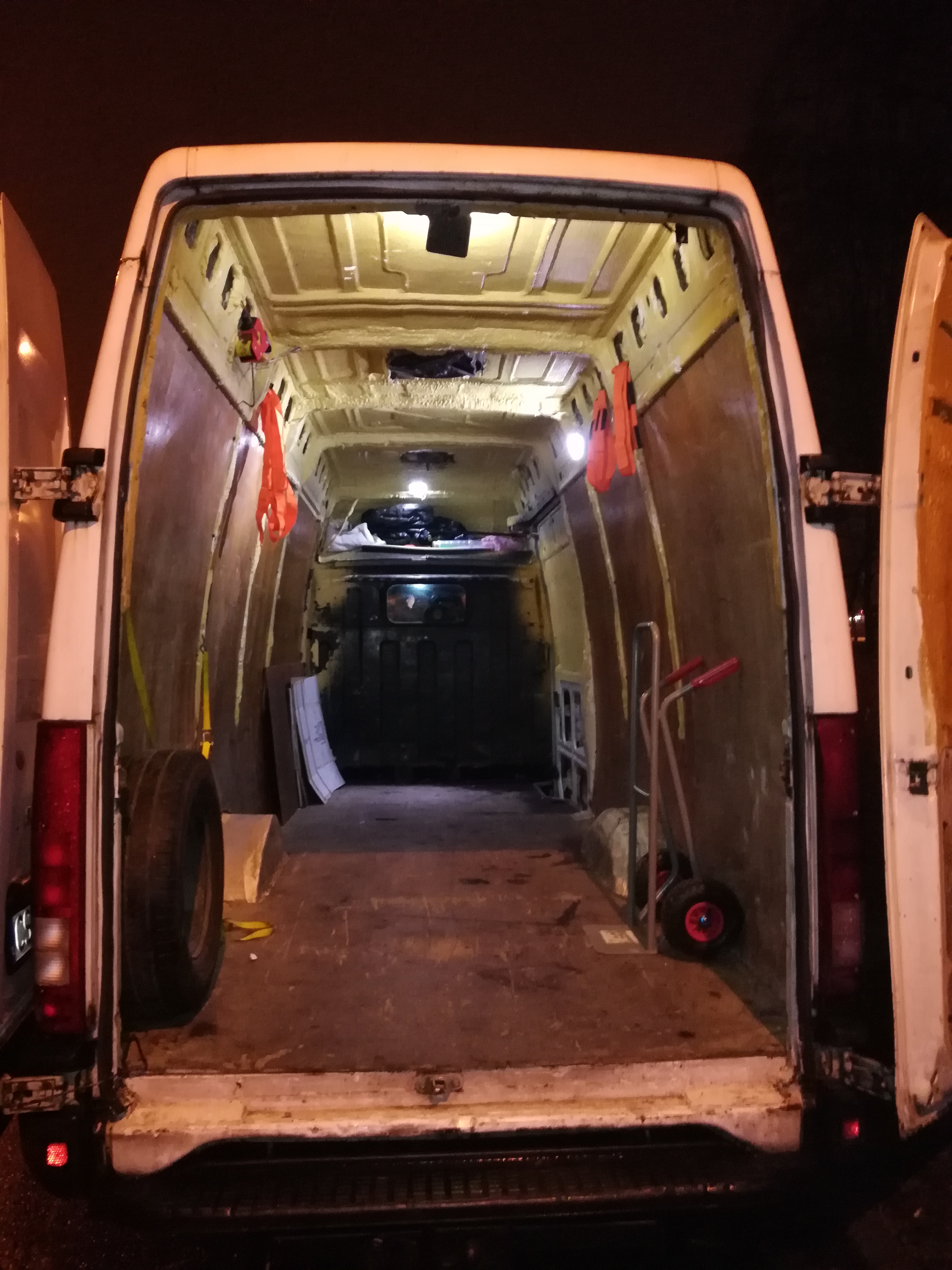 Wnętrze pustego, oświetlonego busa z otwartymi drzwiami, widoczna opona zapasowa, wózek transportowy i zabezpieczenia ładunku.