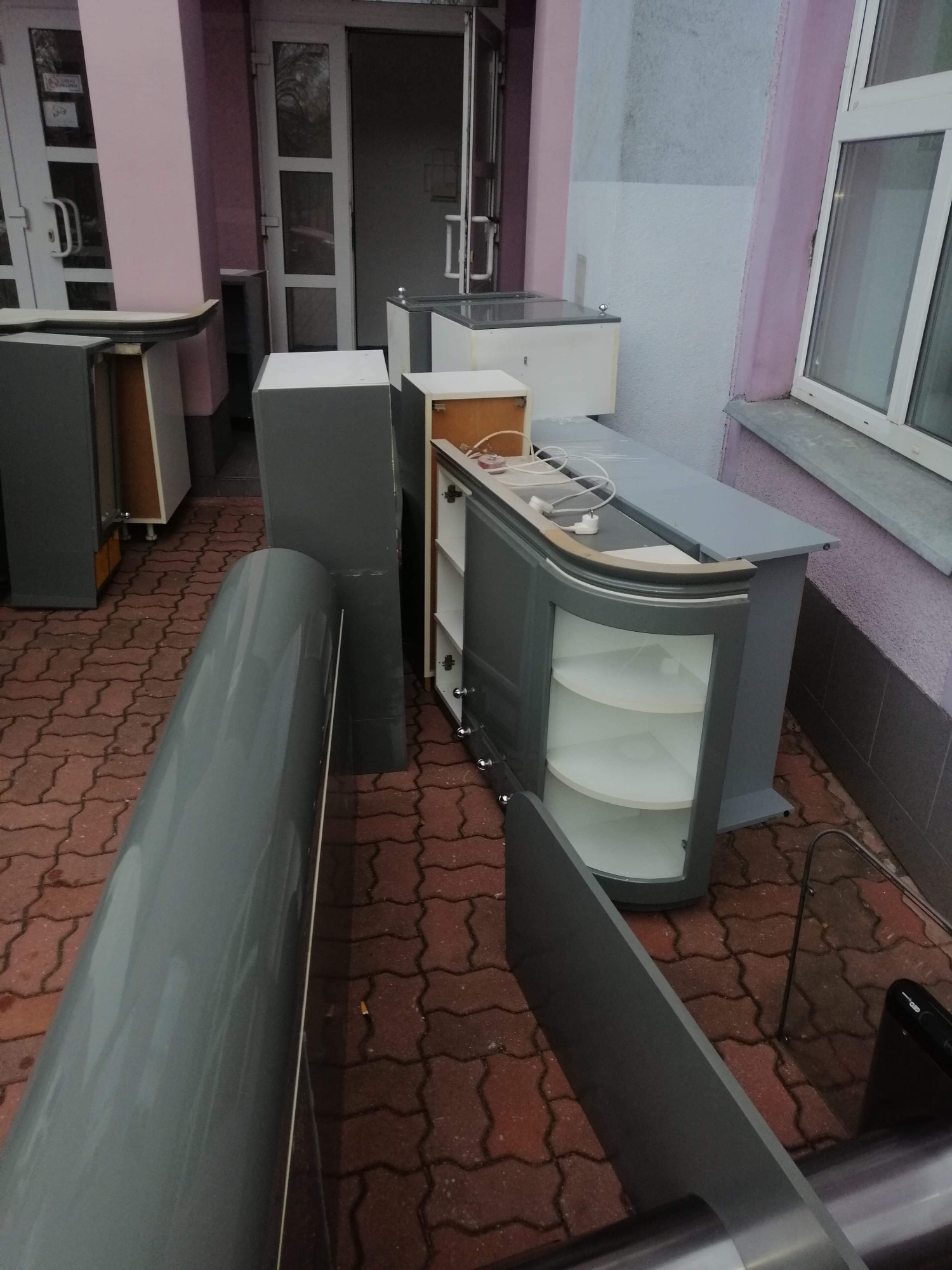 Demontaż i wynoszenie szarych mebli kuchennych z budynku na zewnątrz, widoczne otwarte drzwi i kostka brukowa.