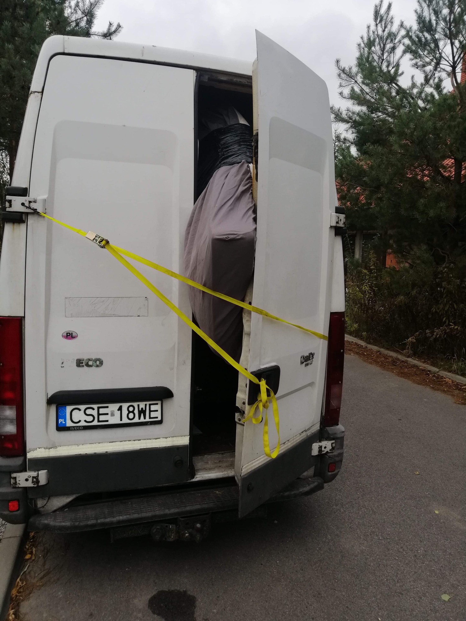 Otwarty tył białego busa Iveco Daily z polską rejestracją CSE 18WE, zabezpieczony żółtymi pasami transportowymi, wewnątrz widoczny mebel owinięty szarą folią ochronną.