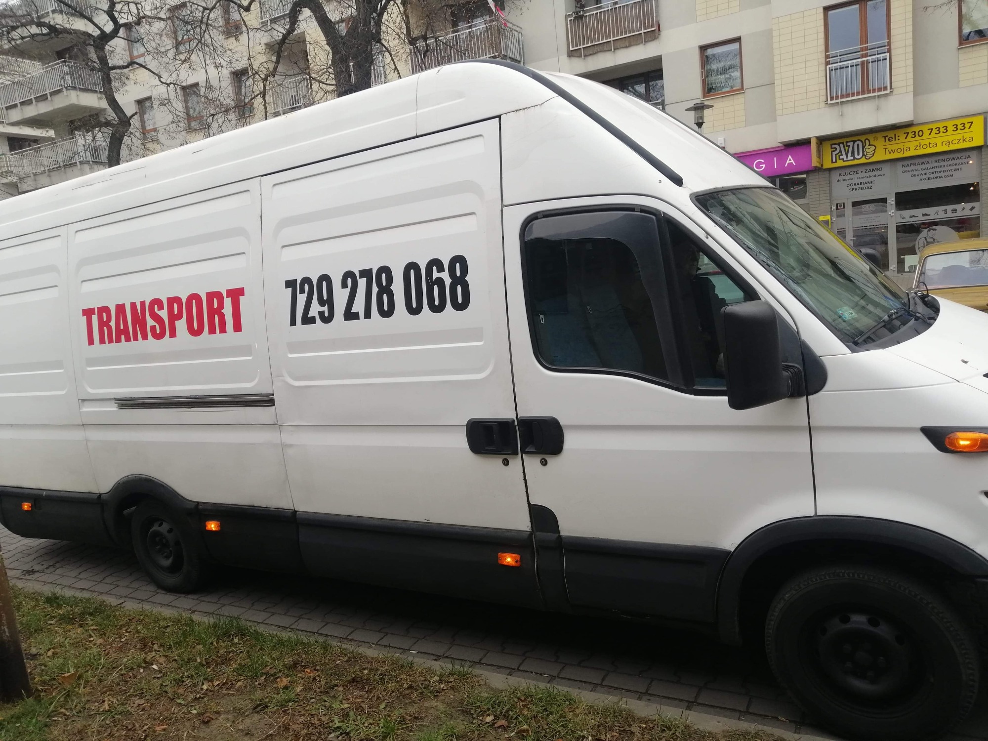 Biały samochód dostawczy z czerwonym napisem TRANSPORT i czarnym numerem telefonu 729 278 068, zaparkowany na ulicy w miejskiej scenerii z budynkami w tle.