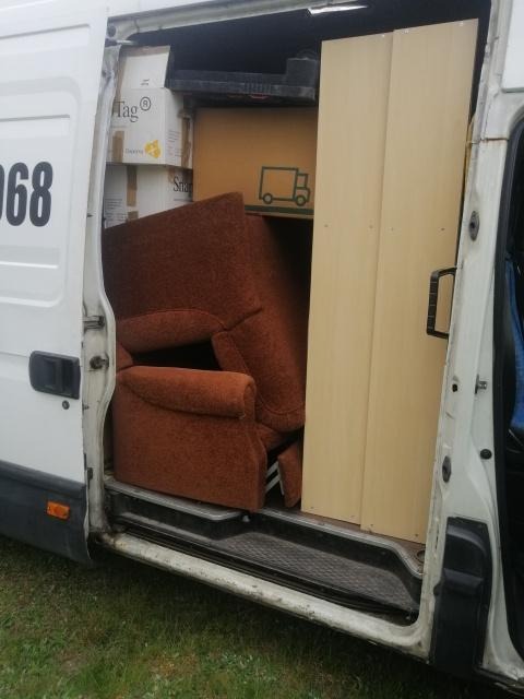 Wnętrze białego busa załadowane kartonami, brązowym fotelem i elementami mebli, widoczne przez otwarte drzwi przesuwne.