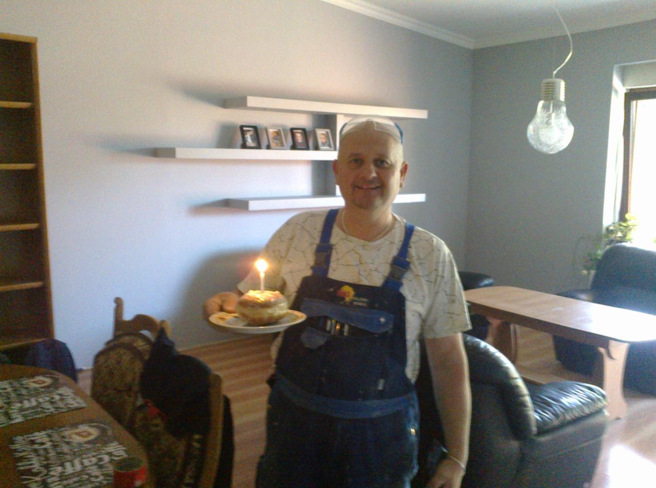 Pracownik w kombinezonie, stojący w pomalowanym na szaro pokoju w Łagowie, trzyma tort ze świeczką, świętując prawdopodobnie zakończenie prac.
