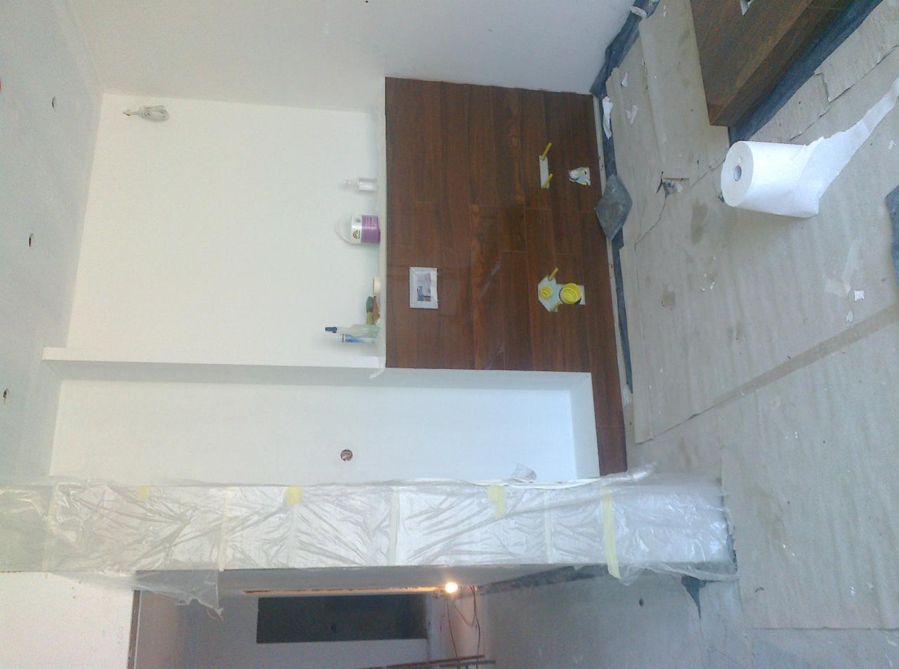 Łazienka w trakcie remontu: ściany częściowo wyłożone brązowymi płytkami imitującymi drewno, białe ściany z półką, zabezpieczona folią sufitowa konstrukcja i rolka papieru na podłodze.