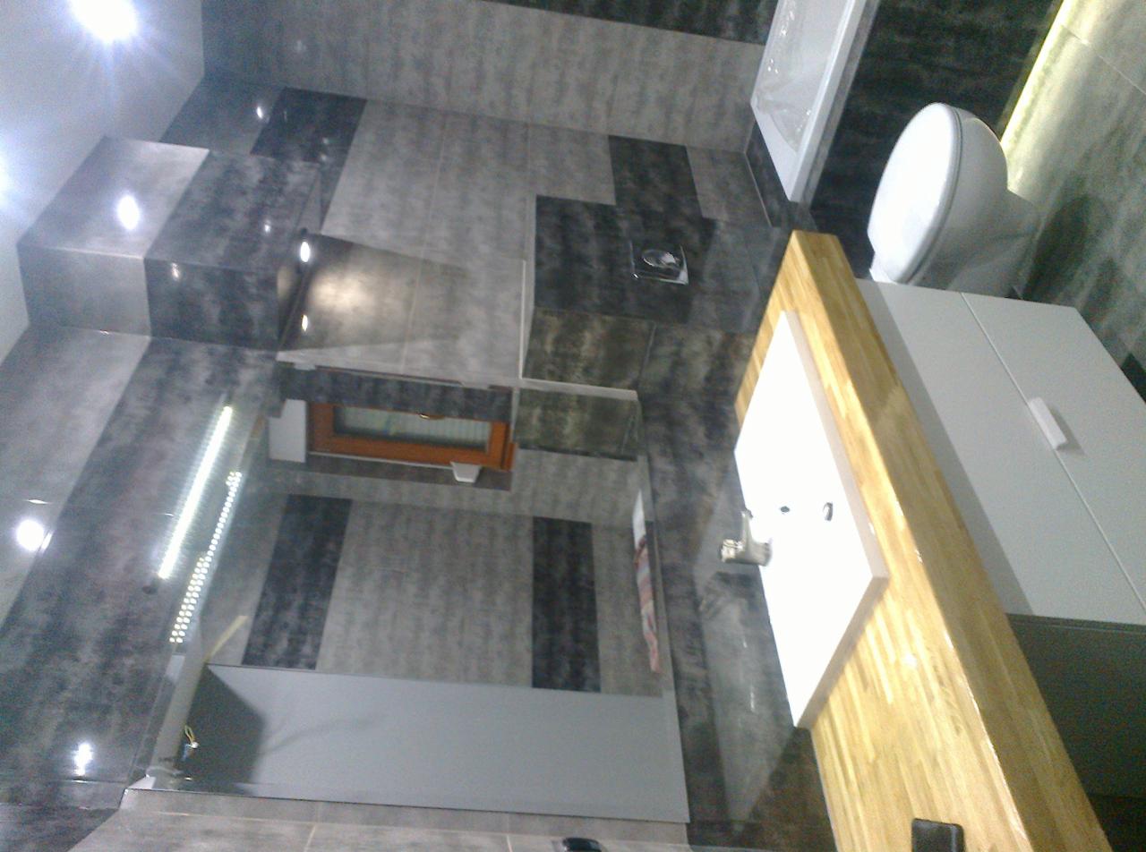 Nowoczesna łazienka z szarymi płytkami imitującymi kamień, oświetleniem punktowym w suficie i drewnianą ramą lustra nad umywalką.