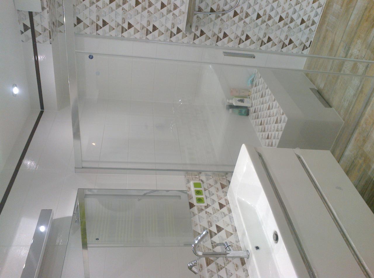 Nowoczesna łazienka z kabiną prysznicową typu walk-in, umywalką z szafką i geometryczną mozaiką na ścianach, widoczny chromowany kran i gniazdko elektryczne z trzema polami.