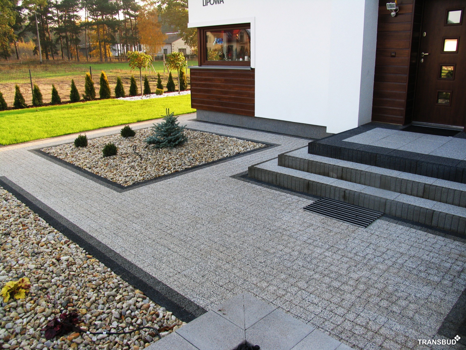 Szara kostka brukowa ułożona przed nowoczesnym domem z drewnianymi elementami, obok rabata z kamieniami i małą choinką, w tle zielony trawnik i drzewa.