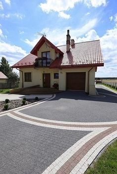 Ułożona kostka brukowa w kolorze grafitowym z obrzeżem z kostki w kolorze beżowym, tworząca półkole przed domem jednorodzinnym z czerwoną dachówką, błękitne niebo z chmurami w tle.