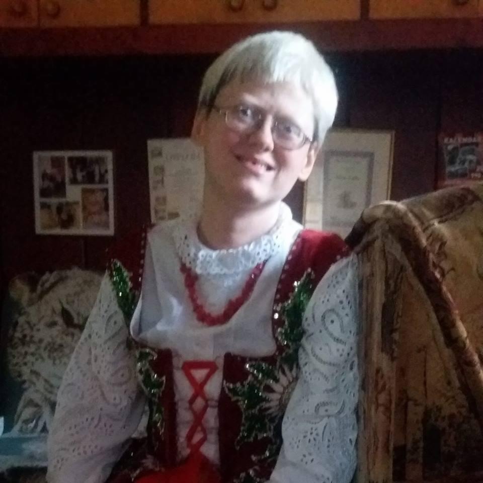 Uśmiechnięta kobieta w okularach, ubrana w tradycyjny strój ludowy z białej koronki, czerwonego aksamitu i zielonych zdobień, pozuje na tle ciemnych mebli i dekoracji.