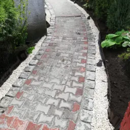 Ścieżka z szarej i czerwonej kostki betonowej ułożona w ogrodzie, obrzeża z jasnych kamyków, roślinność po bokach.