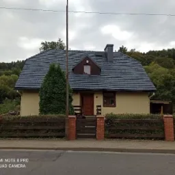 Dom jednorodzinny z ciemnym dachem i jasną elewacją, otoczony drewnianym płotem z ceglanymi słupkami, widok z ulicy.