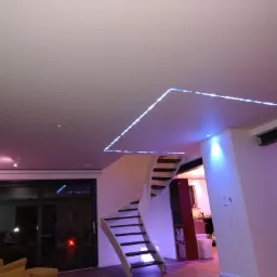 Nowoczesne wnętrze z białymi ścianami i schodami, oświetlone niebieskimi LEDami na suficie, z elementami wystroju w odcieniach różu i żółci.