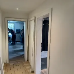 Korytarz w trakcie remontu, widoczne świeżo osadzone, pomalowane na biało ościeżnice drzwiowe, zabezpieczona podłoga i fragment łazienki z kabiną prysznicową w tle