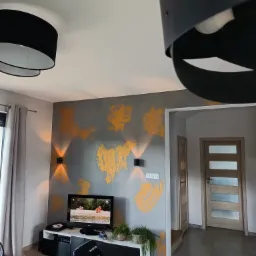 Szara ściana z dekoracyjnymi, pomarańczowymi plamami w nowoczesnym salonie z czarnymi lampami i telewizorem.