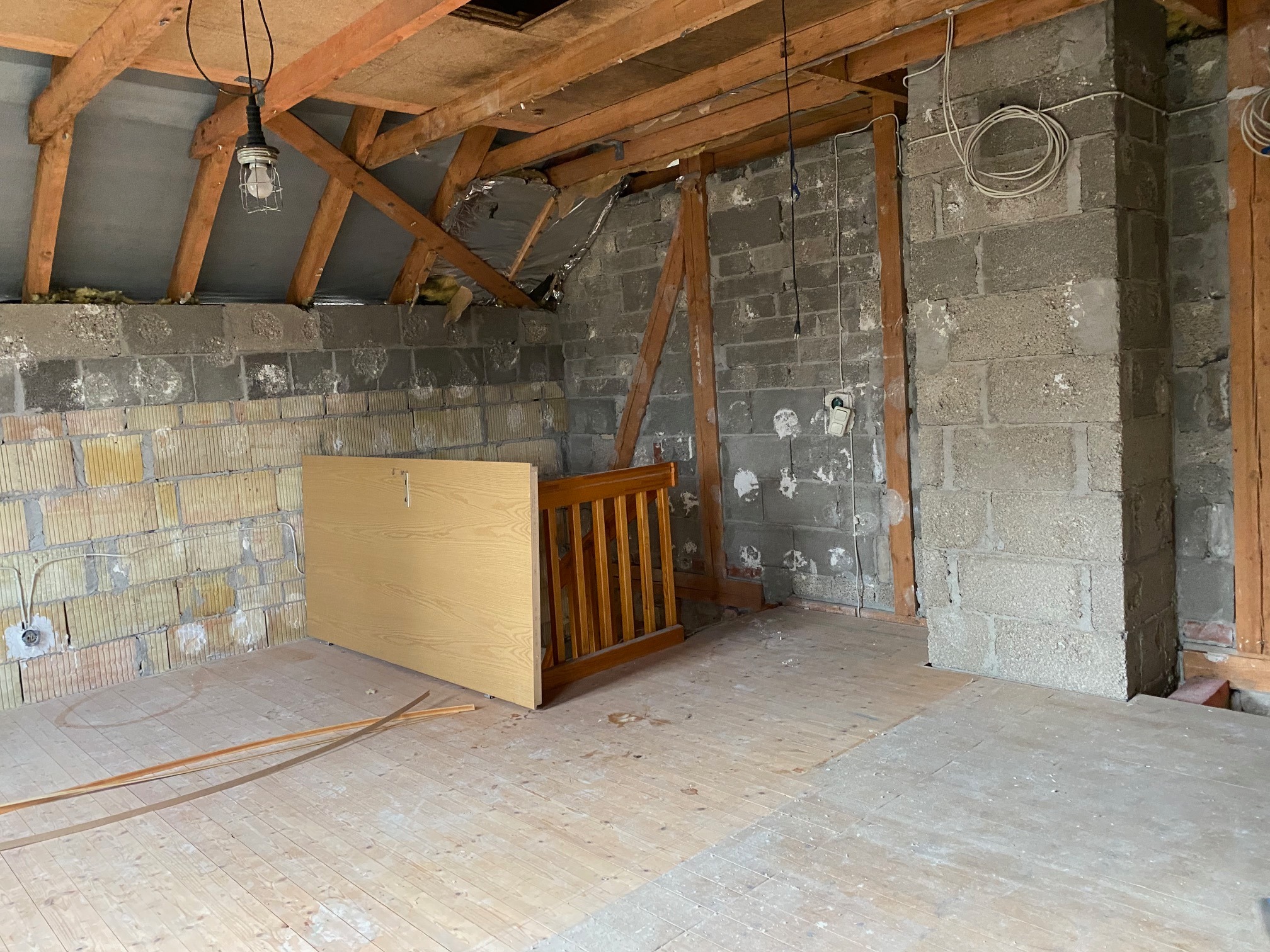Surowe wnętrze poddasza w trakcie remontu: odsłonięte bloczki betonowe ścian, drewniana konstrukcja dachu, prowizoryczne oświetlenie żarówką, fragmenty drewnianej podłogi i element balustrady.