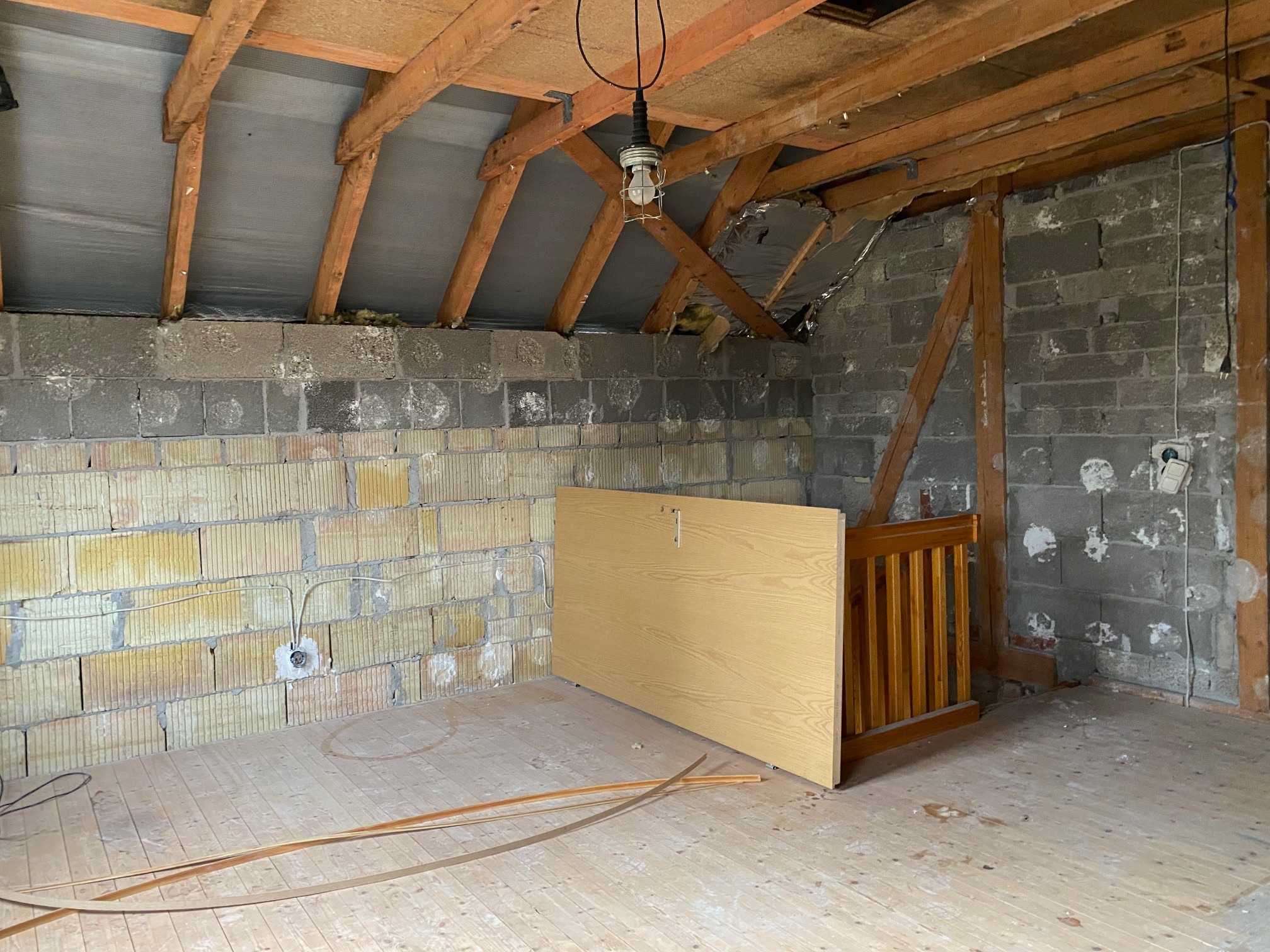 Adaptacja poddasza: surowe ściany z bloczków, odsłonięta konstrukcja dachu z widocznymi belkami, drewniana podłoga w trakcie remontu, wisząca lampa budowlana i oparte o ścianę elementy drewniane.