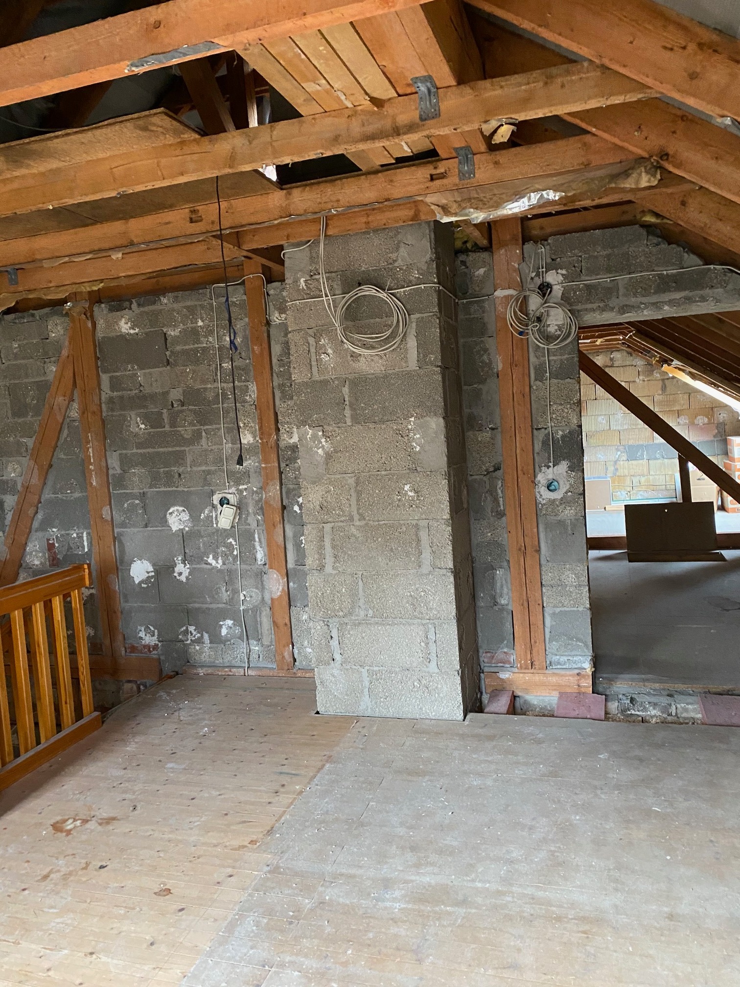 Poddasze w trakcie remontu: surowe ściany z bloczków, drewniana konstrukcja dachu, widoczne przewody elektryczne i częściowo położona drewniana podłoga.