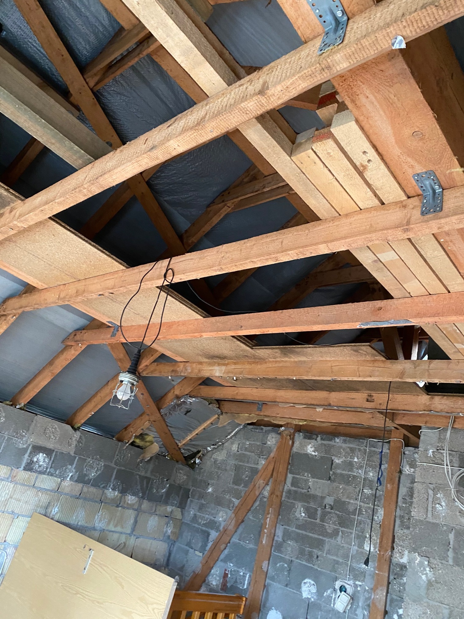 Drewniana konstrukcja dachu od wewnątrz, z widocznymi elementami izolacji i prowizorycznym oświetleniem w trakcie prac remontowych.