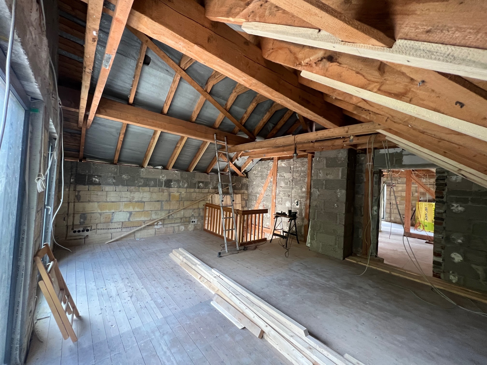 Poddasze w trakcie adaptacji: surowe ściany z bloczków, drewniana konstrukcja dachu z widoczną izolacją, drewniana podłoga, przygotowane deski, drabina oparta o balustradę, widoczne kable elektryczne.