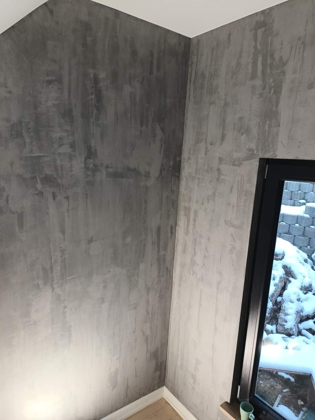 Ściana wewnętrzna wykończona szarą, dekoracyjną gładzią w stylu betonu architektonicznego, widok na okno z widokiem na ośnieżony ogród.