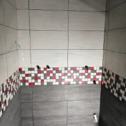 Świeżo wykafelkowana ściana łazienki z mozaiką w czerwono-biało-szare kwadraty, widoczne wystające rury instalacji hydraulicznej i niedokończony sufit podwieszany.
