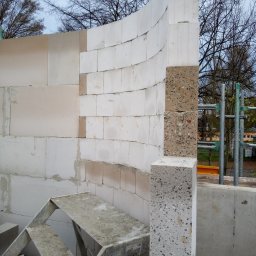 F.B.K.G - Wznoszenie ściany z białych bloczków, z fragmentem łuku i słupem z betonu lastryko. Widoczne rusztowanie i drabina. Praca w toku, surowy stan.