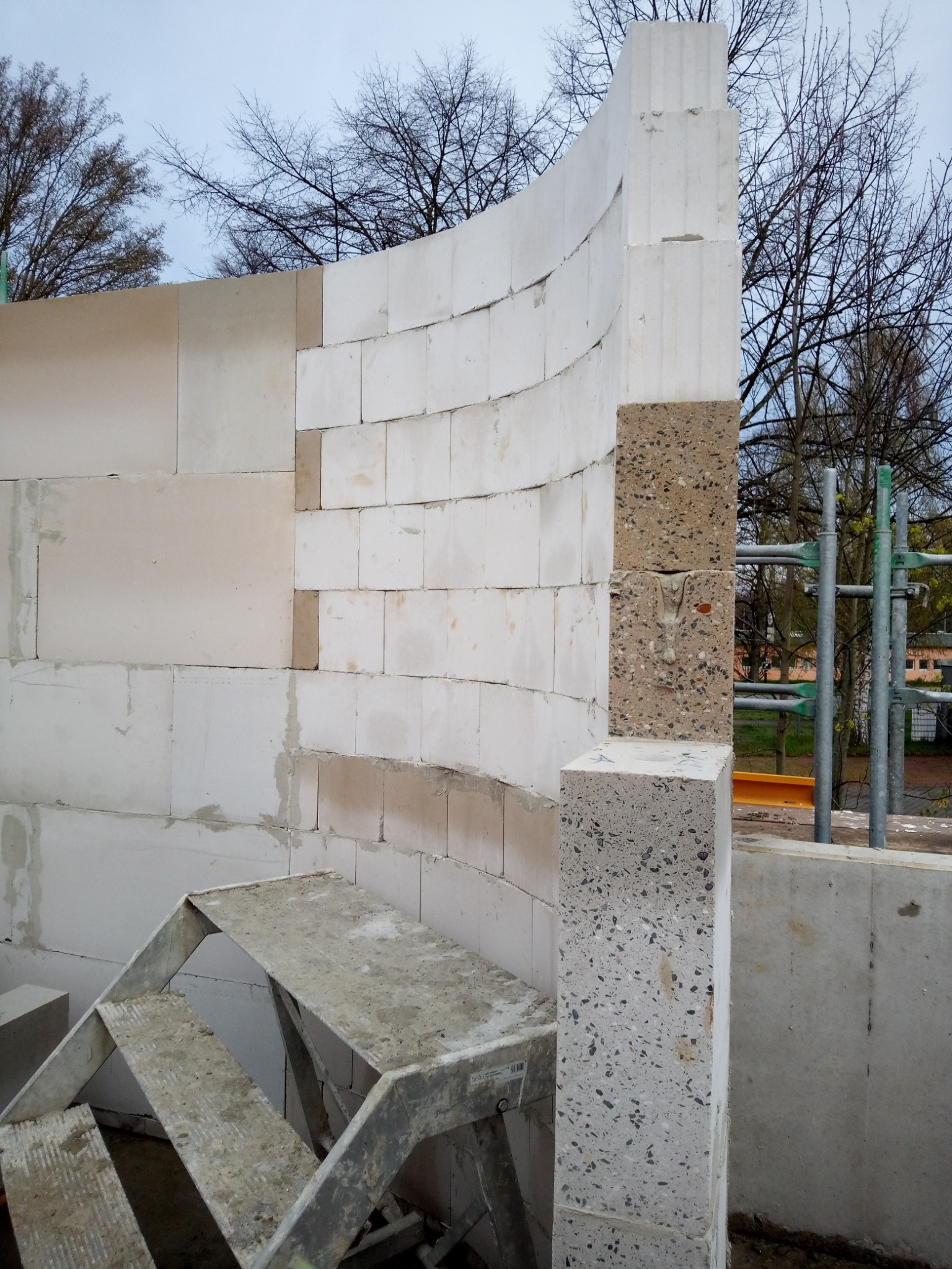 Wznoszenie ściany z białych bloczków, z fragmentem łuku i słupem z betonu lastryko. Widoczne rusztowanie i drabina. Praca w toku, surowy stan.
