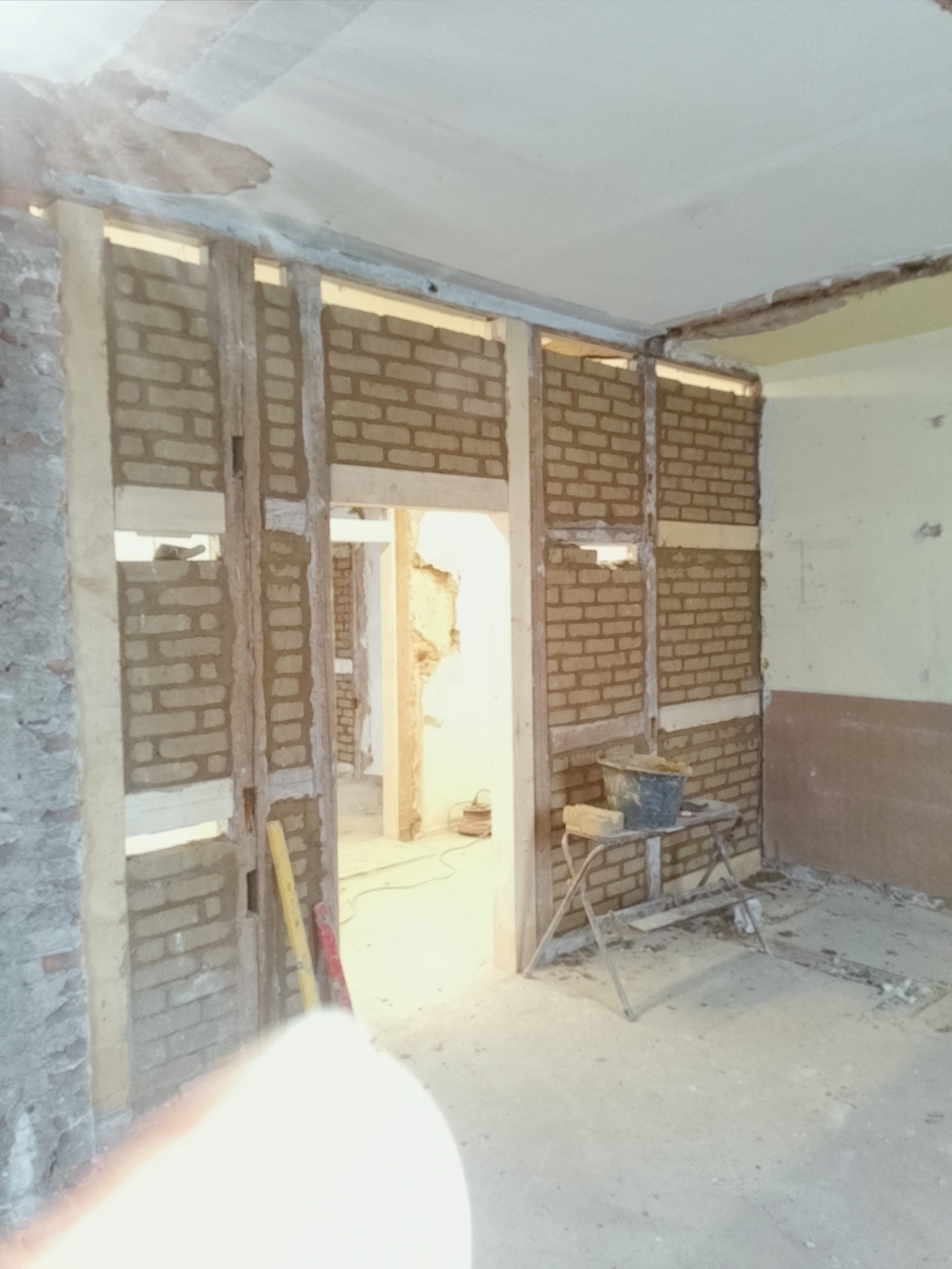 Wnętrze w trakcie murowania ścian z cegły, widoczne drewniane elementy konstrukcyjne, narzędzia murarskie i fragmenty starego tynku. Prace remontowe w pomieszczeniu.