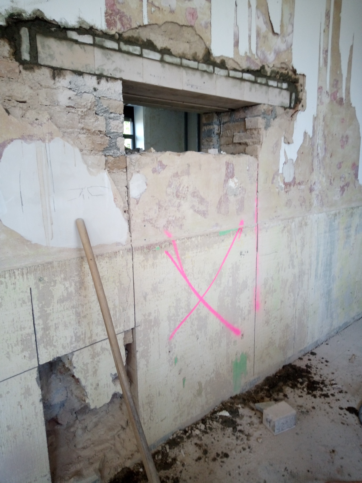 Fragment ściany w trakcie remontu z odsłoniętą konstrukcją, widoczne ślady po kuciu i różowe oznaczenia na ścianie. W tle okno.