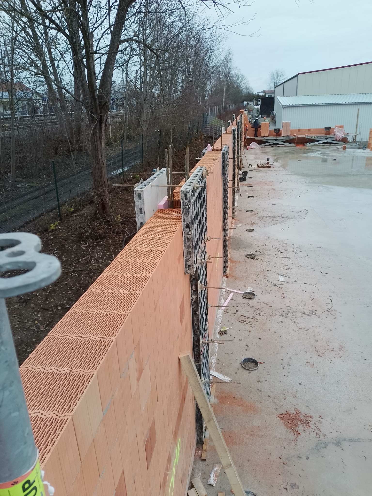 Wznoszenie ściany z pustaków ceramicznych Porotherm z użyciem szalunków systemowych. Widoczne zbrojenia i elementy mocujące. Tło: drzewa, budynek przemysłowy.