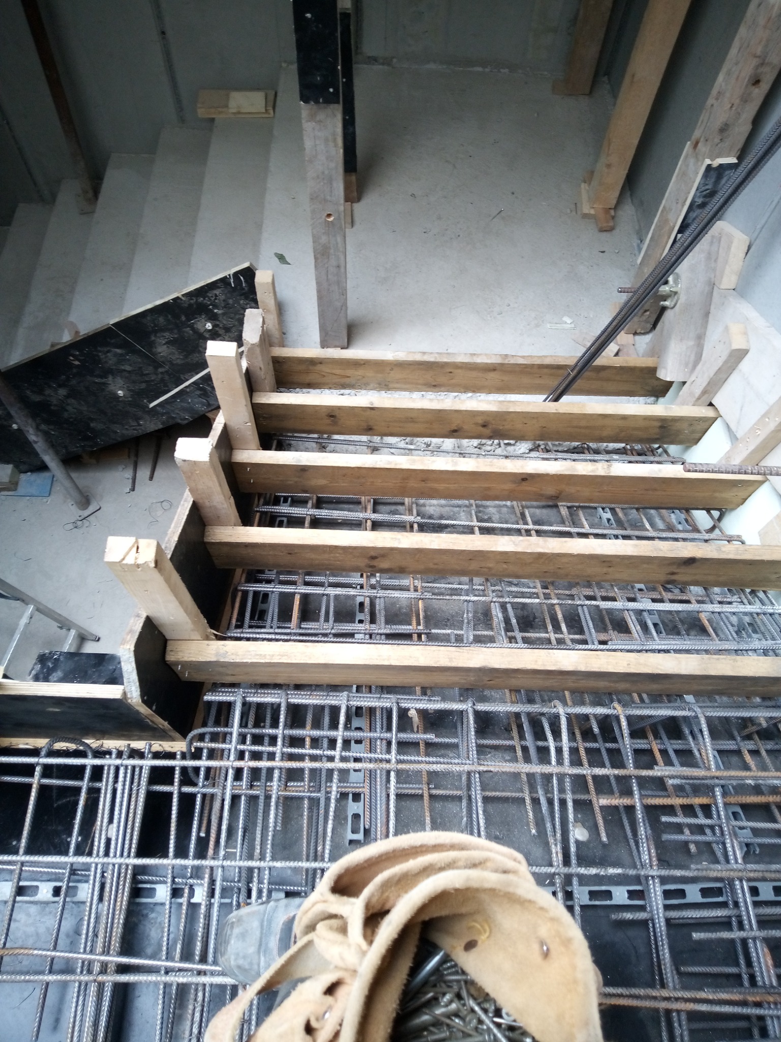 Zbrojenie schodów z drewnianym szalunkiem, widok z góry. Metalowe pręty tworzą siatkę, a drewniane elementy podtrzymują konstrukcję. W tle widoczne betonowe schody.