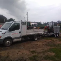 Biała ciężarówka z platformą załadowana minikoparką i traktorem, ciągnąca przyczepę z kolejną minikoparką na błotnistym terenie.