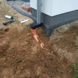 Instalacja rury odprowadzającej wodę deszczową przy fundamencie budynku, widoczna wykopana ziemia, narzędzia i fragment budynku z izolacją.