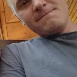 Mężczyzna robiący selfie z drewnianą konstrukcją w tle. Ujęcie z dołu, widoczna część szarej koszulki i fragment dłoni.