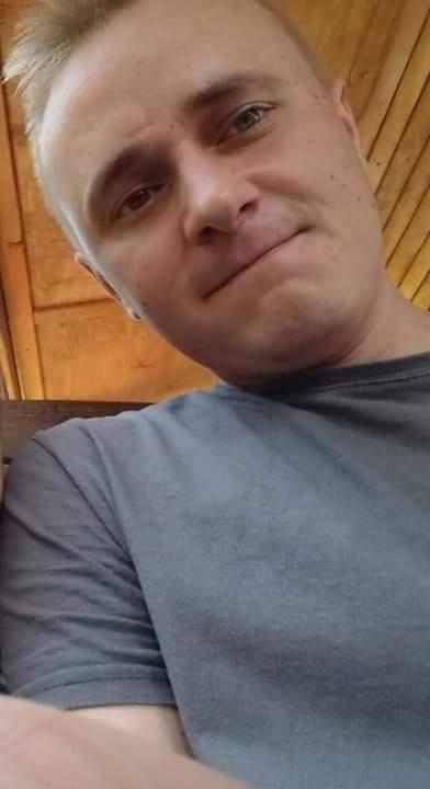 Mężczyzna robiący selfie z drewnianą konstrukcją w tle. Ujęcie z dołu, widoczna część szarej koszulki i fragment dłoni.