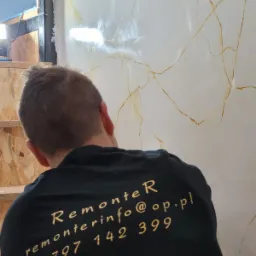 Pracownik w czarnej koszulce z napisem 'remonterinfo@op.pl 797 142 399' stoi tyłem do kamery, wykonując dekoracyjną strukturę na ścianie imitującą marmur z wyraźnymi złotymi żyłkami.