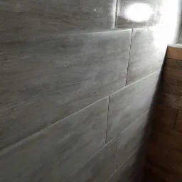 Ściana wyłożona szarymi, podłużnymi płytkami imitującymi drewno, połączona z fragmentem ściany wyłożonej brązowymi, drewnianymi panelami.