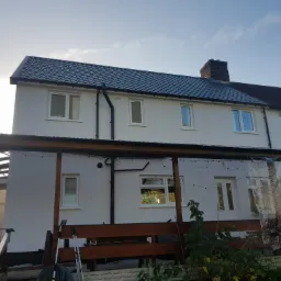 Odnowiona elewacja dwupiętrowego domu z nowym dachem z blachodachówki, widoczna z poziomu tarasu z drewnianą balustradą i rozwieszonymi lampkami.