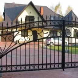 Czarna, ozdobna brama wjazdowa z kutego żelaza na tle domu jednorodzinnego z brązowym dachem i podjazdem z czerwonej kostki brukowej. Widoczne elementy dekoracyjne w postaci zawijasów i grotów.