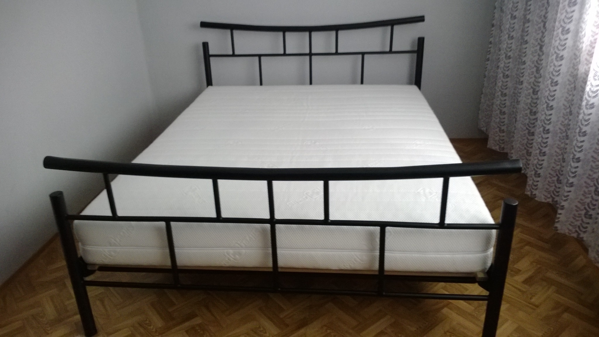 Czarne metalowe łóżko z materacem w minimalistycznym wnętrzu z drewnianą podłogą i zasłoną w tle.