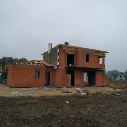 Dwukondygnacyjny dom w trakcie budowy z czerwonej cegły, widoczne elementy konstrukcji dachu, szary krajobraz w tle.