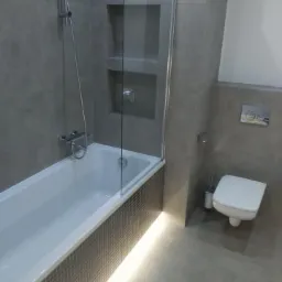 Nowoczesna łazienka w szarościach z podświetlaną wanną obudowaną mozaiką, szklanym parawanem nawannowym i wnęką prysznicową w ścianie.