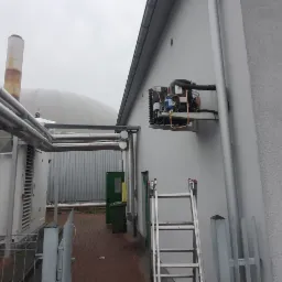 Agregat chłodniczy zamontowany na zewnętrznej ścianie budynku, widoczna drabina oparta o ścianę, w tle instalacja przemysłowa i zbiornik biogazu.